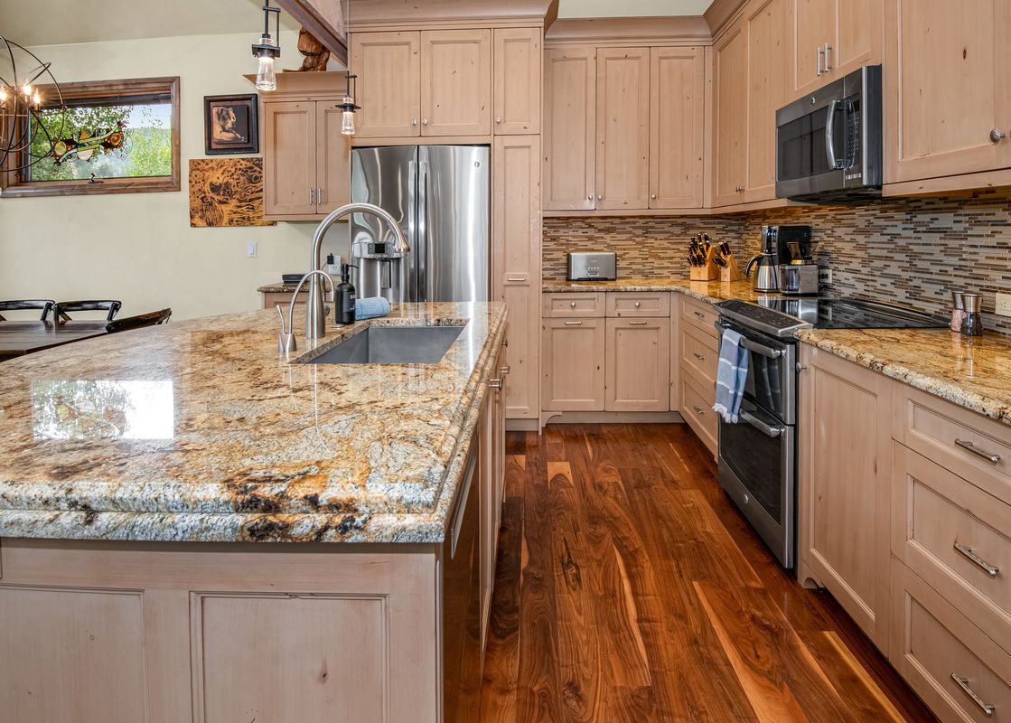 A modern kitchen with granite countertops, light wood cabinetry, hardwood floors, stainless steel appliances, and a window with a view of green trees.