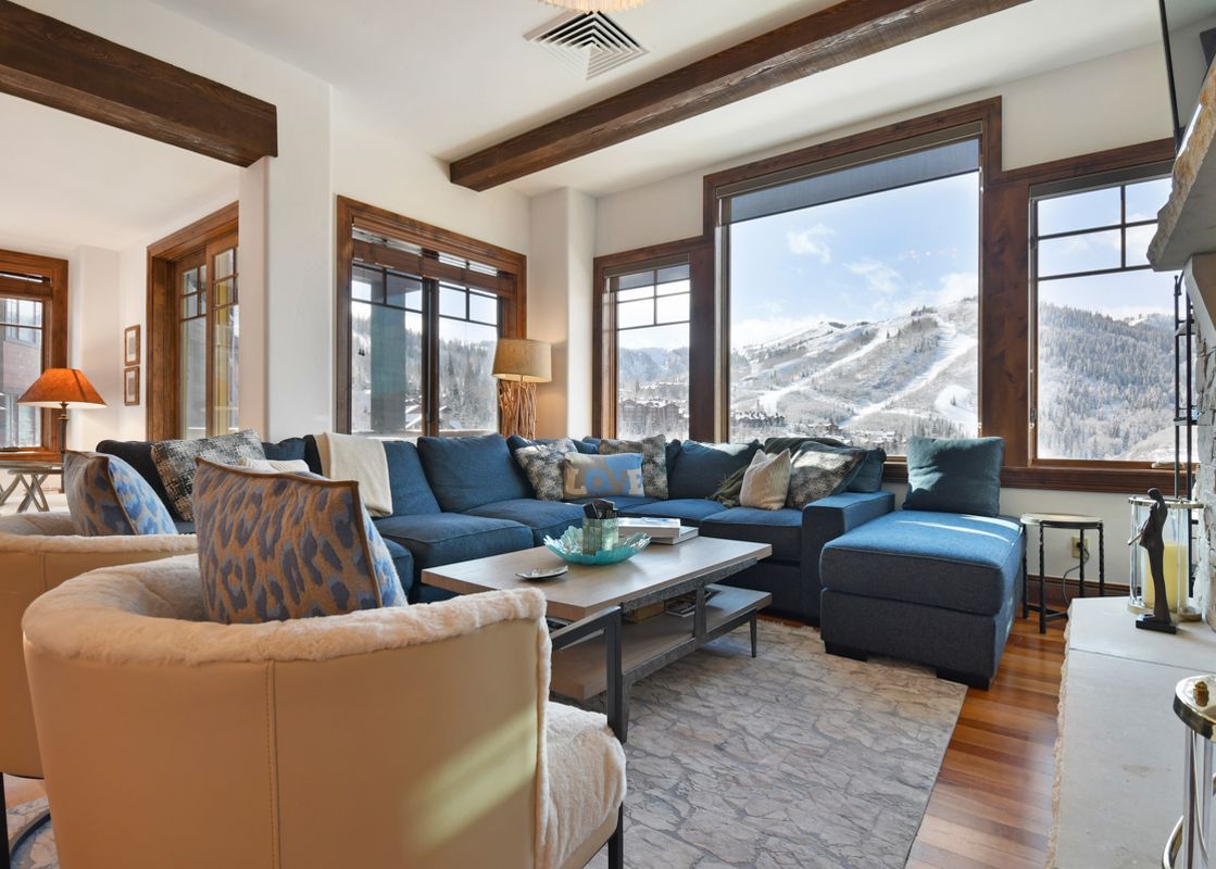 The living area of Silver Strike Lodge 605, featuring a large blue sectional sofa, a light-colored coffee table, and two cream armchairs, with a stunning view of snow-covered mountains through large windows.