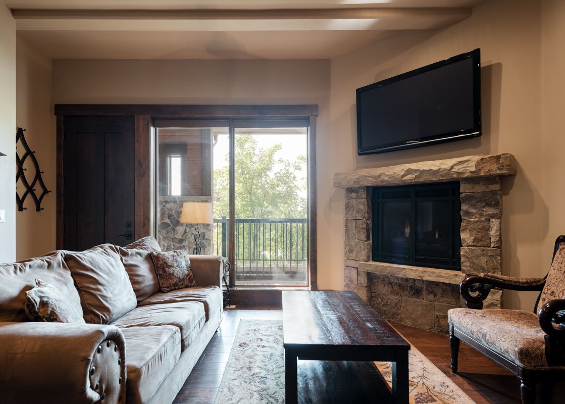 A cozy living room at 103 Juniper Landing features a large sectional sofa, a wooden coffee table, a stone fireplace with a TV, and a glass door leading to a balcony.