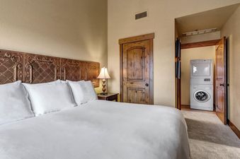 A cozy bedroom with a white bed and an ornate headboard, adjacent to a wooden door that reveals a stacked washer and dryer unit.