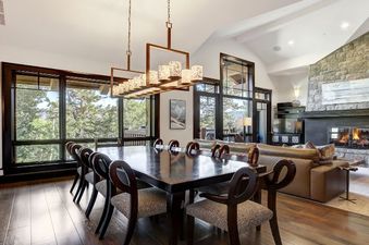 A wide-angle shot of the dining and living area in the Hummingbird Platinum 3 BR Plus Loft, featuring a large dining table with elegant chairs, a modern chandelier, a stone fireplace, and large windows with an outdoor view.