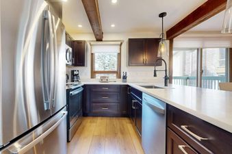 A modern kitchen with stainless steel appliances, dark wood cabinets, light countertops, and a double window with a view to the outside.