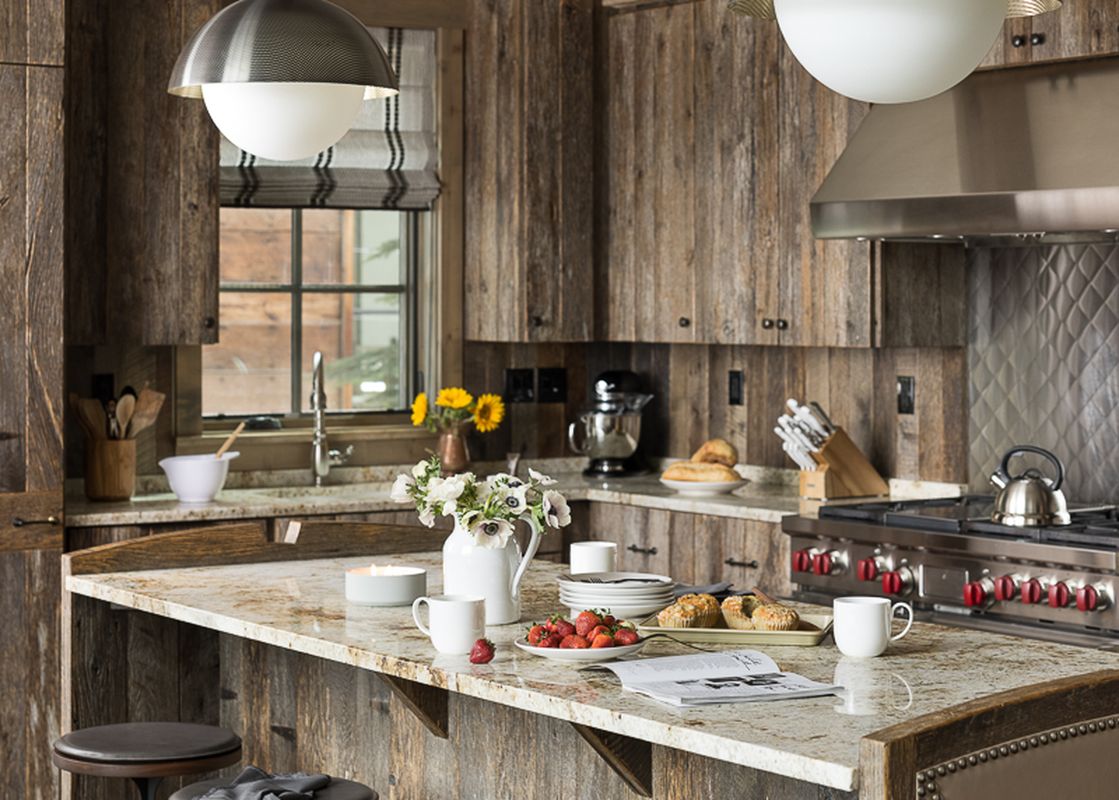 A rustic-chic kitchen at Fish Creek Lodge 05 features reclaimed wood cabinetry, a granite island with breakfast items, and modern stainless steel appliances.