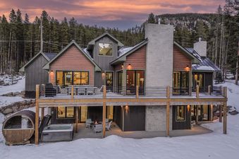 A stunning winter evening view of the Everwild Chalet, showcasing its illuminated interior and exterior, a large deck, a hot tub, and a fire pit amidst a snowy forest backdrop with a dramatic sunset sky.