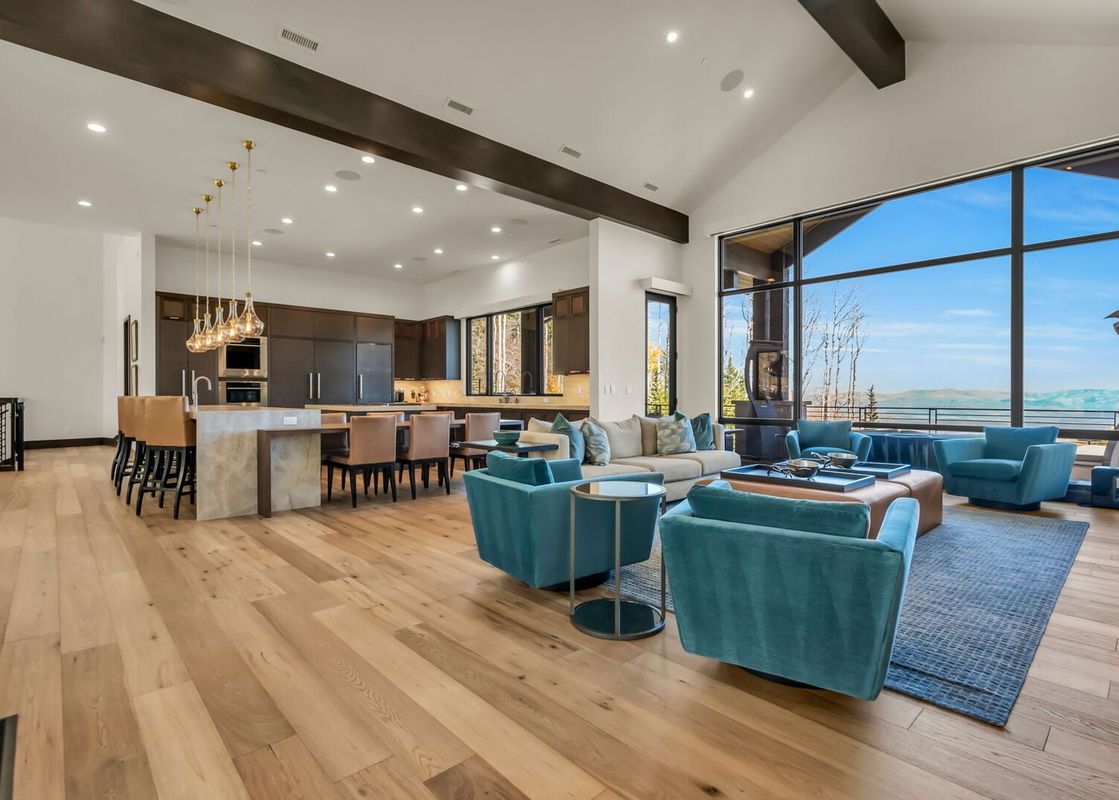 A wide shot of the open-concept living area and kitchen at Sanctuary Chalet at The Colony, featuring blue armchairs, a sofa, and a modern kitchen with a large island and dining area, all with expansive windows offering mountain views.