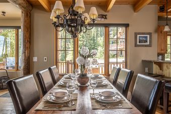 A formal dining table set for six with elegant place settings, a central floral arrangement, and a grand chandelier, all illuminated by natural light from large windows showcasing a vibrant outdoor view of green trees.