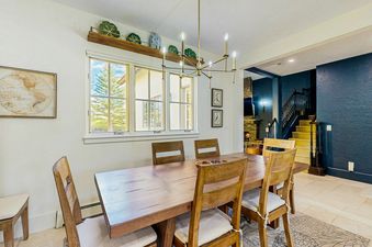 A spacious dining area with a long wooden table, chairs, a decorative shelf, framed artwork, and a large window offering an exterior view of trees.
