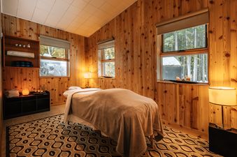 A massage table sits in the center of a room with a wood paneled wall. The room is lit by lamps and features a rug on the floor.