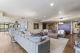A spacious, open-concept living room and kitchen area with light wood floors, gray couches, a modern kitchen with a large island, and a dining area with a mountain view.
