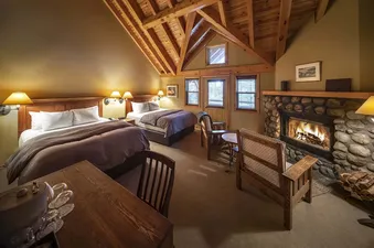 A rustic hotel room with two beds, a stone fireplace, and exposed wooden beams on the ceiling.
