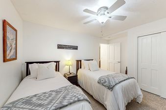 The guest bedroom at Emerald Pines at Motherlode C features two twin beds, a ceiling fan, and a small lamp on a bedside table. The walls are white with artwork, and the carpet is a light brown.