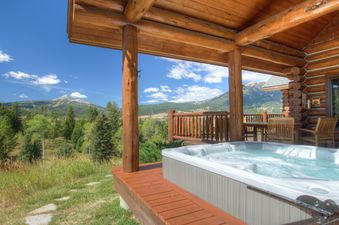A spacious wooden deck at Huckleberry Hideaway features a hot tub, a dining area, and stunning mountain and forest views under a clear blue sky.