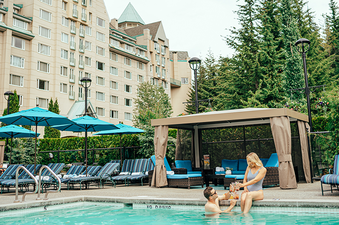 A couple relaxes poolside with drinks in a lush outdoor setting next to the Fairmont Chateu 1 Bedroom Slopeside View King Suite.