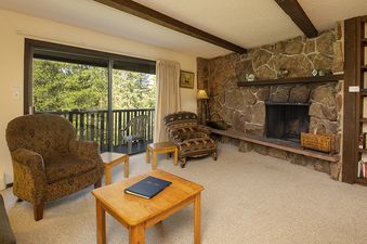 A cozy lounge area in Hi Country Haus 2111 with a stone fireplace, comfortable armchairs, and a sliding glass door leading to a balcony overlooking lush green trees.