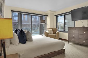 A spacious king bedroom at Lowell 203 featuring a comfortable bed, large windows with an exterior view, a cozy armchair, and a flat-screen TV.
