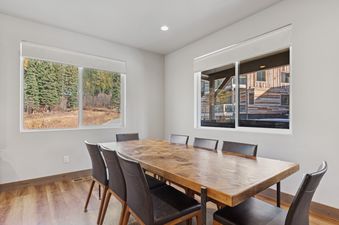 A bright dining area at Sweet Peaks with a large wooden table and dark leather chairs, offering views of autumn foliage and other buildings through two wide windows.