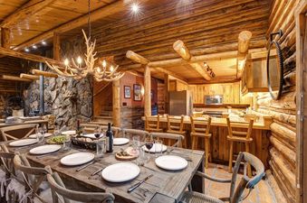 A rustic dining room with a wooden table and chairs, an antler chandelier, and a view of a kitchen.