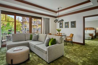A spacious living area with a large sectional sofa, an oval ottoman, and a dining area, featuring a large window with a view of lush greenery and distant buildings, with a partial view of a bedroom through an open doorway.