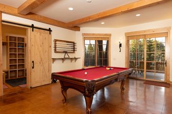 A spacious game room at Fox Paw Lodge features a red-felt pool table, wooden beams, barn doors, and large windows with forest views.