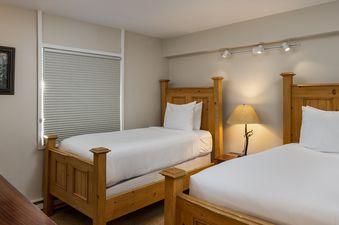 A cozy bedroom at Vantage Point 206 with two twin beds featuring wooden headboards and white bedding. A nightstand with a lamp sits between the beds, and a window with blinds is on the left.