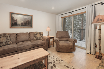The living area at Beaver Village 1612 features a comfortable brown sofa, a matching armchair, a rustic wooden coffee table, and a unique log-style floor lamp, with a large sliding glass door revealing a snow-covered outdoor balcony.