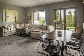 The interior of the Yeti Ridge Retreat features a spacious living room with a large L-shaped sectional sofa and a dark wooden dining table, all illuminated by natural light from windows overlooking a forested mountain view.
