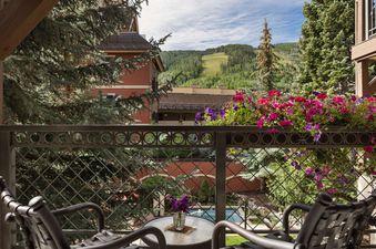 A private balcony at Montaneros 211 offers a scenic view of green mountains, red buildings, and a distant pool, framed by a decorative railing and colorful flowers.