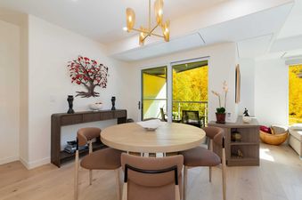 A dining area in Signature Suite 552 with a round wooden table, four brown chairs, a modern chandelier, and a sliding glass door revealing a vibrant yellow autumn landscape outside.