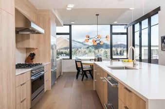 A kitchen with white countertops, wooden cabinets, and a stainless-steel stove and oven. There is a view of the mountains through the large windows behind the kitchen island.