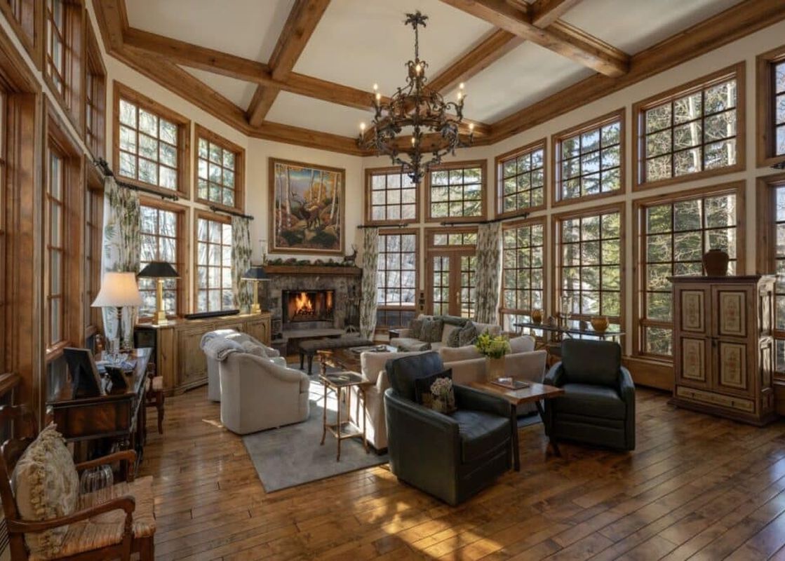 A spacious living area with hardwood floors, a large stone fireplace, and numerous windows offering views of snow-covered trees.