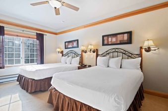 A hotel room at The Charter G140 features two queen-sized beds with white linens, wrought iron headboards, bedside tables with lamps, framed art, a large window, and a ceiling fan.