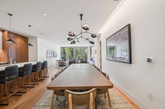 A modern dining area with a long wooden table and stylish chairs, connected to a kitchen with dark wood cabinets and a living room with large windows.