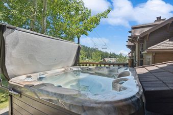 A private hot tub with its cover partially open, showing clear water, set on a deck with lush trees, a ski lift, and a building in the background under a blue sky.
