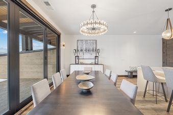 A modern dining room features a long dark wood table surrounded by gray upholstered chairs, illuminated by a contemporary circular chandelier, with a large glass door revealing an outdoor patio.
