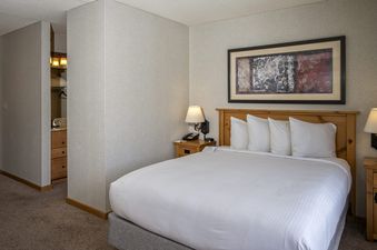 A comfortable bedroom with a queen-sized bed, white linens, wooden headboard, bedside tables with lamps, and framed artwork, leading into a vanity area.