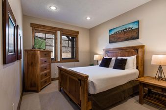 A cozy guest bedroom at Town Pointe C304 features a wooden bed with white linens and dark pillows, a wooden dresser with a TV, and two windows with outdoor views.