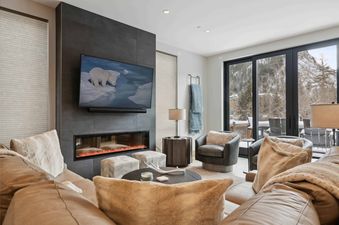The inviting lounge area at One Riverfront Townhome 05 features a large tan leather sectional, a sleek linear fireplace, and a flat-screen TV, all set against a stunning snowy mountain backdrop visible through expansive glass doors.