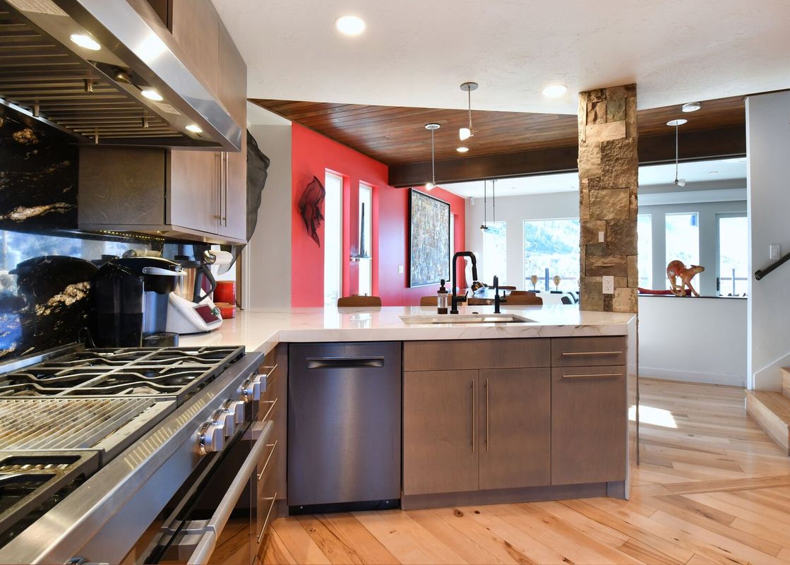 A modern kitchen with stainless steel appliances, dark wood cabinets, and a white marble countertop, featuring a black granite backsplash and an open concept view to a living area with red accent walls.