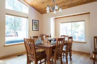 The dining area at North Shore Golden Pine features a wooden table set with dishes and wine, surrounded by chairs, with large windows overlooking a scenic outdoor view and a hot tub.