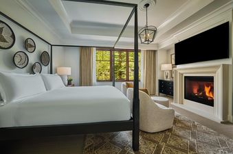 A large, bright bedroom features a king-sized bed with white linens, a black four-poster frame, and decorative woven baskets on the wall.