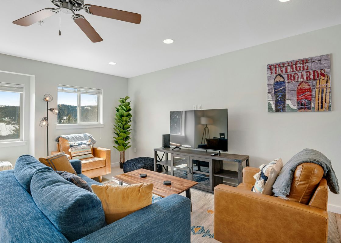 A cozy living room featuring a blue sofa, brown leather armchairs, a wooden coffee table, a flat-screen TV on a media console, and large windows with a view of a snowy landscape.