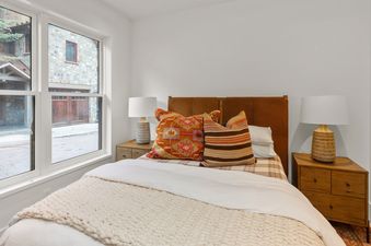 A cozy bedroom at Depot Place features a bed with a chunky knit throw, patterned pillows, a leather headboard, and wooden nightstands with lamps next to a window.