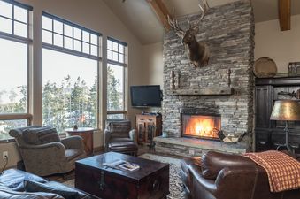 A cozy living room in Wolf Pack Hideaway with a large stone fireplace, comfortable seating, and expansive windows showcasing a winter landscape.