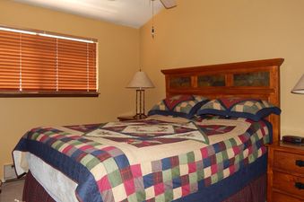 The bedroom in Beaver Village 131 features a wooden headboard, a colorful patchwork quilt, and a window with blinds.