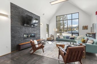 A modern alpine living room featuring a stacked stone fireplace, large windows with mountain views, a teal sectional sofa, and stylish wooden armchairs.