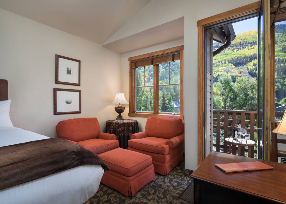 A cozy bedroom with a bed, two orange armchairs, and a window and balcony overlooking a scenic mountain view.