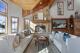 A cozy living room in Bellemont 5 with a stone fireplace, large TV, and plush white sofas, offering mountain views through multiple windows.