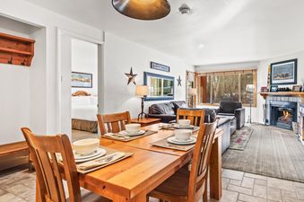 A dining table set for four in a cozy living area with a fireplace and large windows showcasing a winter scene.