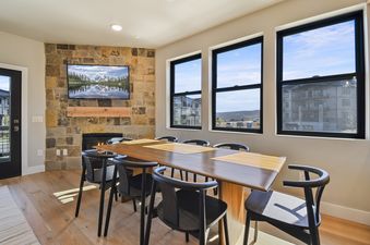 A dining area at Mayflower Lakeside B5 104 featuring a long wooden table, black chairs, a stone accent wall with a mounted TV, and a fireplace, with large windows overlooking an exterior view.