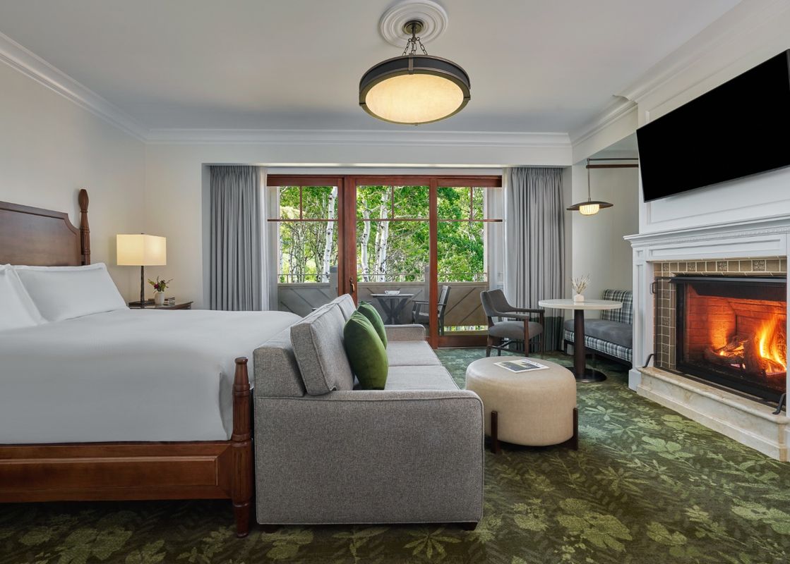 The Montage Deer Valley King Room Resort View features a luxurious king-sized bed, a comfortable sofa, and a cozy fireplace, all set against a backdrop of natural beauty visible through large windows leading to a private balcony.